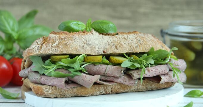 Woman adding basil to delicious sandwich with pastrami (cured beef meat), pickles and arugula at white table, closeup