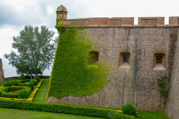 Historic stone fortress wall with ivy and watchtower overlooking lush gardens and distant ocean, manicured gardens with geometric hedges are visible below, leading to a distant view of the ocean © Pixel-Shot