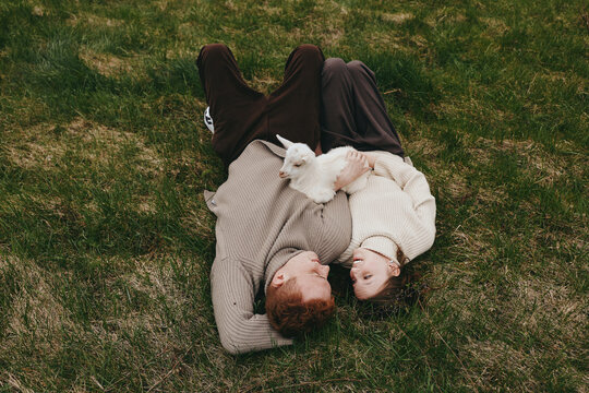 A couple lies on a grassy field, cuddling a white kid goat between them, sharing a calm moment in the open air. A gentle, lifestyle scene focused on family bonding and rural simplicity.