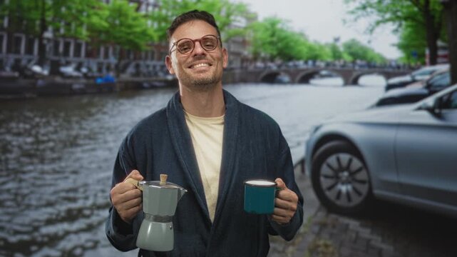 Man in robe holding a moka pot in left hand and a blue cup in right hand, smiling with visible hands on a canal street beside parked cars and a bridge; contentment.