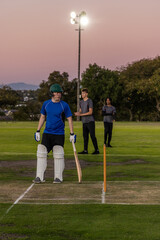 Obraz premium Diverse male teammates standing on cricket pitch at dusk, batter holding wooden bat near stumps