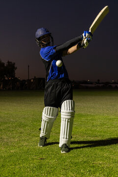 Cricket batter is swinging wooden bat with white ball on grass field at dusk under lights