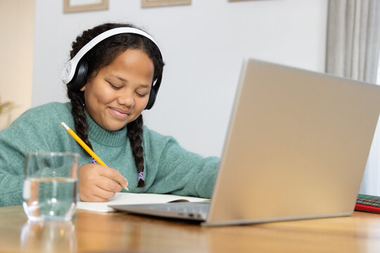 Female child writing with yellow pencil at wooden table at home, wearing headphones, laptop nearby