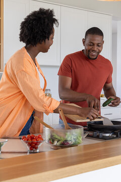 African American couple cooking together in kitchen, making salad bowl with cucumber and frying pan