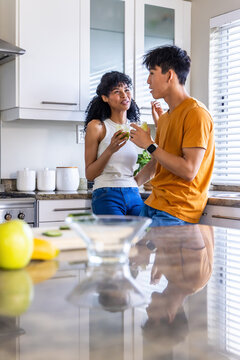 Diverse couple chatting on modern kitchen countertop, holding green drinks near fruit bowl