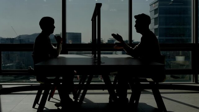 Silhouettes of two people sitting across from each other and having an animated conversation with hand gestures at a desk in a dark moody office with a bright city skyline visible through large window