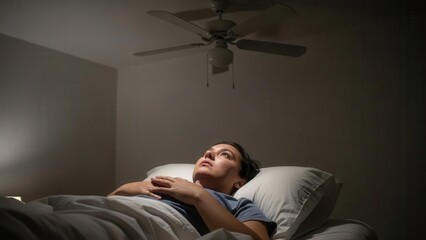Fototapeta premium A woman lies awake in bed at night, looking up at the ceiling fan in a dimly lit room, unable to sleep.