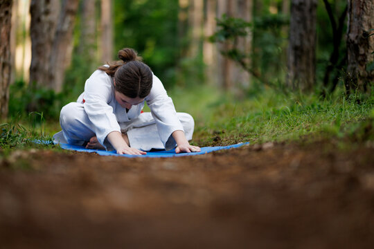 Young woman in white karate gi stretching on a blue mat during outdoor martial arts training in a peaceful forest