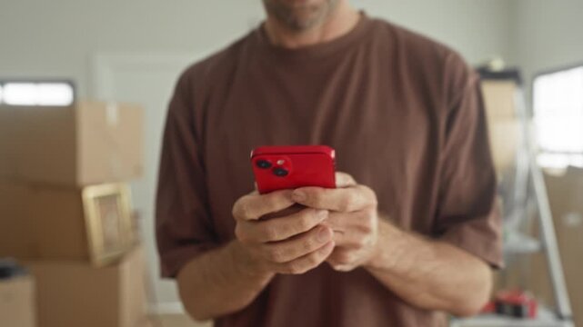 Man holding red smartphone, looking at phone and gesturing amid packed moving boxes inside a building; moving frustration.