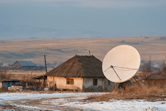 Snow-dusted field holds rustic cabin with thatched roof. Large satellite dish stands beside the weathered house. Winter light casts soft hues over distant hills and sparse trees