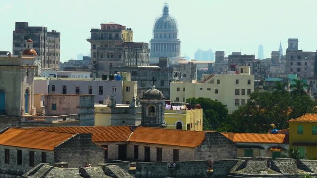 Aerial perspective slowly moving across the weathered rooftops and colorful buildings of old Havana, showing the iconic dome of El Capitolio Nacional against the hazy skyline on a sunny day