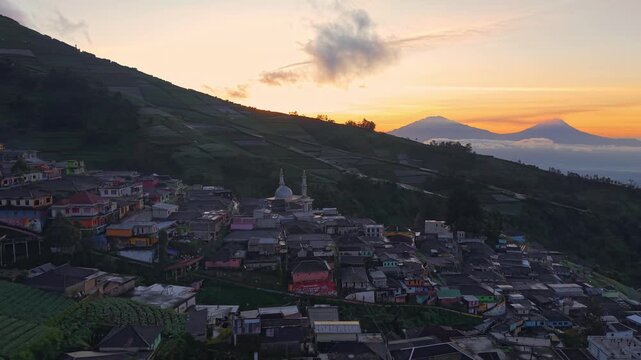 Aerial view of Scenic mountain village at sunrise with colorful sky, rural homes, and misty hills. Peaceful countryside landscape in tropical highlands captured from above. Nepal Van Java, Indonesia.