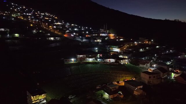 Elevated night aerial view of a village with bright lights and winding streets. Rural development, evening atmosphere, and scenic landscape. Nepal Van Java, Indonesia.