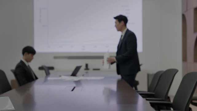 A businessman in a dark suit standing beside a projection screen displaying a line chart while a colleague sits at a large conference table during a business meeting in a modern boardroom.