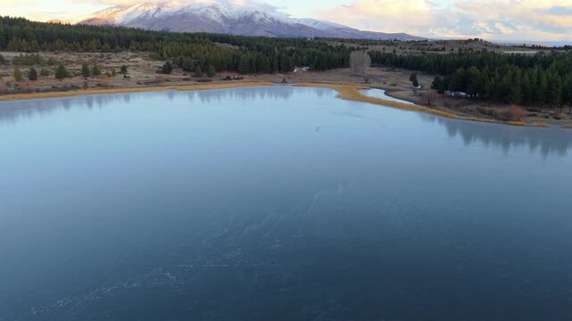 Drone lateral slide above Laguna La Zeta near Esquel, Chubut, Argentina, revealing calm alpine lake, Patagonian forest, shoreline wetlands, and snow covered Andes.
