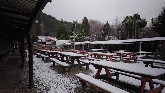 Quiet winter scene showing rows of empty wooden picnic tables covered in fresh snow at an outdoor market in Colonia Suiza, Argentina.