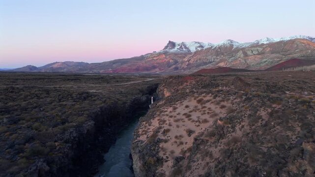 Stunning drone footage of a river flowing through a dark volcanic canyon at sunrise in La Payunia Reserve, Mendoza, Argentina. Beautiful pink sky.