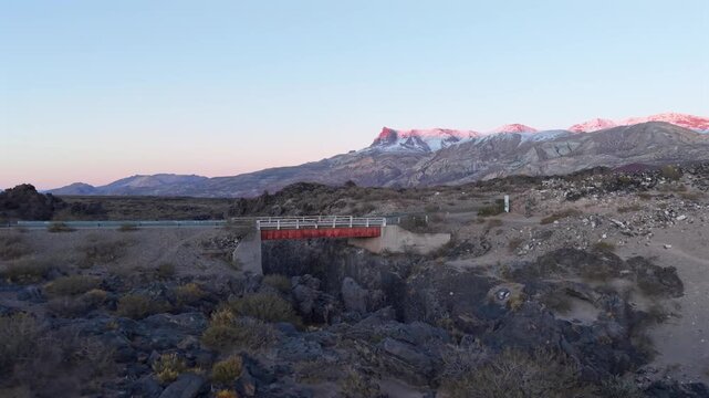 Drone footage of a red bridge spanning a dark volcanic canyon in La Payunia Reserve, Mendoza, Argentina. Snow-capped mountains glow in the background.