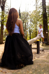 Fototapeta premium Child and Woman Playing on a Wooden See Saw in a Sunlit Park Surrounded by Trees