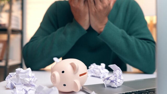 A stressed person crumpling paper with a fallen piggy bank, symbolizing debt burden and financial failure, while also reflecting frustration, lack of savings stability, and emotional strain.
