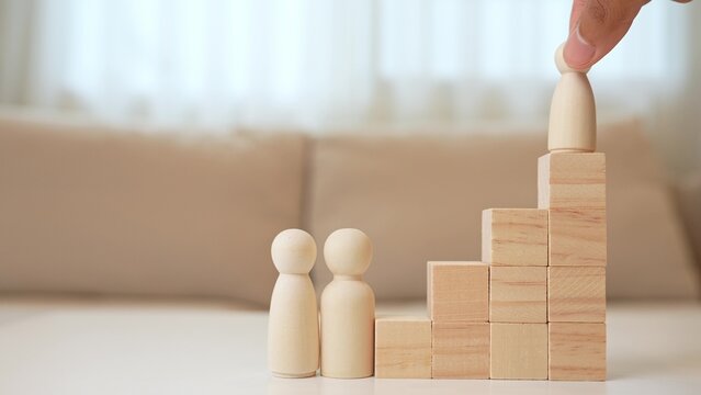 A hand moves a wooden child figure up block steps to the top, beside parent figures, representing growth, encouragement, achievement, parental support, guidance, aspiration, and family success.