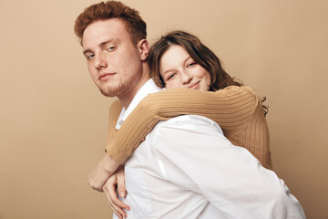 Fototapeta premium Young couple in casual clothes embraces against beige background. Woman hugs man from behind with smile, man looks confident. Concept of love, relationship, affection, and togetherness.