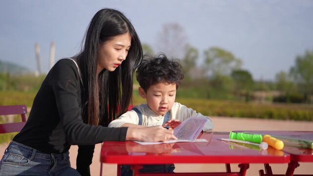 Mother guides child at red picnic table. Child draws with crayon, focused and curious. Outdoor setting with soft sunlight, green trees behind. Scene captures quiet, nurturing learning moment