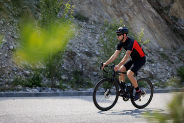 Fototapeta premium Cyclist rides along mountain road in daylight near rocky terrain with trees in background