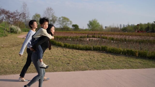 Sunlit park path with three people walking together. Man carries child on shoulders, woman walks beside them. Everyone smiles, enjoying the outdoors together