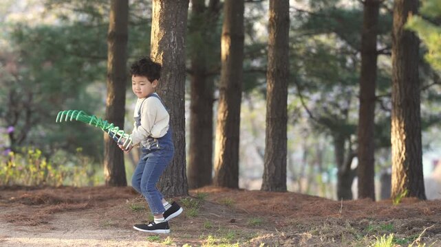 Child plays with rake in wooded area. Bright light filters through tall trees. Natural setting invites calm, playful energy. Perfect for nature, childhood, outdoor activity themes