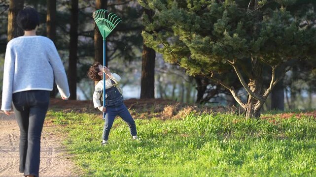 Little child plays joyfully with a rake in a park. Adult watches nearby, smiling gently. Sunlight bathes grass and trees warmly. Child laughs, swinging the rake with delight
