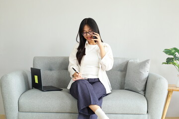 Obraz premium A young woman is working on a laptop, talking on the phone while taking notes in a book, busy working at home, sitting on the sofa at home, the concept of working from home.