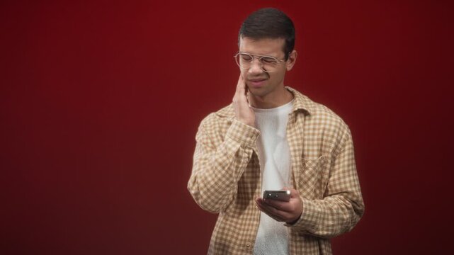 Man holding smartphone touches his neck with one hand and winces while reading a message in studio, wearing glasses and a checkered shirt; discomfort.