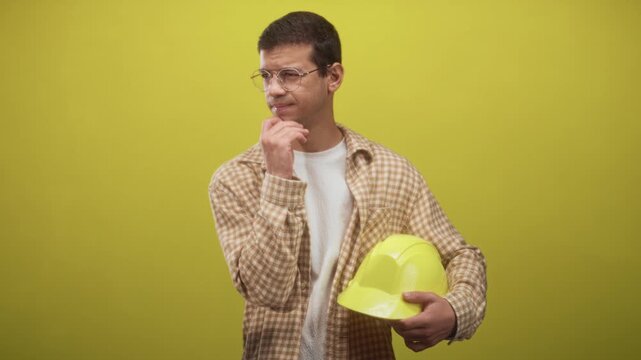 Man holding yellow hardhat, wearing plaid shirt and glasses, with hand on chin in studio; thoughtful.