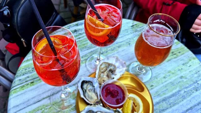 Aperol spritz cocktails, beer and fresh oysters served on a caf&eacute; table, creating a colorful outdoor dining and seafood aperitif scene.