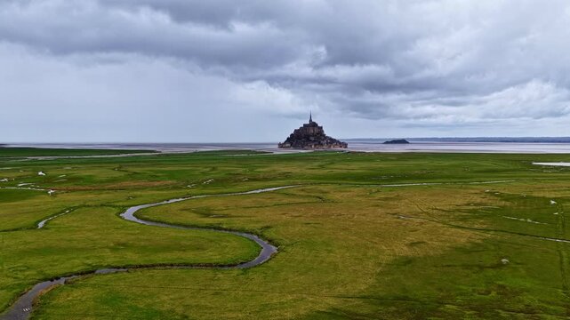 Flowing streams meander through green marshlands towards Mont Saint-Michel, set against a dramatic cloudy sky. Ideal for travel features or historical documentaries.