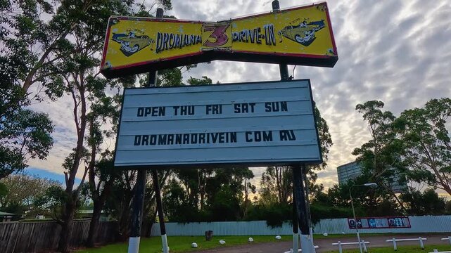 The Dromana 3 Drive-In sign displays opening days and website info, set against a backdrop of trees and a cloudy sky.
