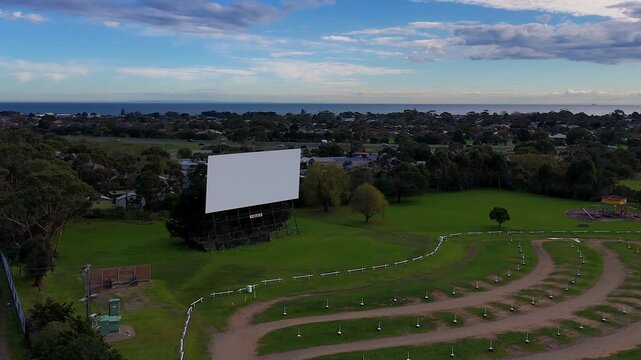 An elevated view of a drive-in theater screen set against a coastal town and ocean backdrop.