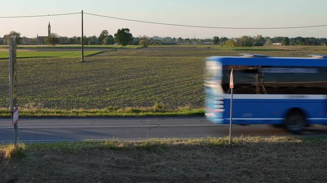 Static Shot of Seta Bus and Cars Speeding Along SS10 Rural Road With Agricultural Fields and Church Tower in Castelvetro Piacentino Portraying the Rhythm of Everyday Life in Emilia Romagna