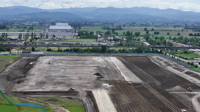 Large scale logistics and datacenter construction site in the Po Valley, Italy, with Frecciarossa high speed rail traffic moving toward Bologna beside highway infrastructure and agricultural plains