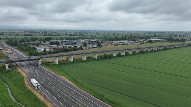 Aerial drone panning right over A21 and A1 motorway junction with SS9 Via Emilia trucks and railway viaduct crossing green Po Valley fields in Fiorenzuola d'Arda Emilia Romagna Italy