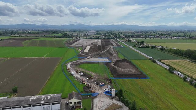 Logistics park construction with earthworks and machinery near A1 motorway and SS9 Via Emilia, Po Valley, Northern Apennines background. Wide drone ascending establishing shot