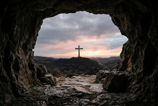 View from inside a dark rocky tomb looking at a wooden cross on a distant hill at sunset
