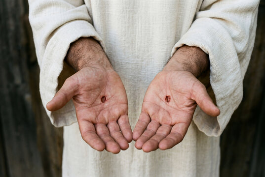 Open palms of a man wearing a bright tunic revealing bloody stigmata wounds indicating sacrifice