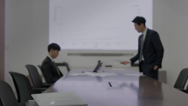A businessman in a dark suit leaning over a conference table to review documents while a colleague sits nearby with a projection screen displaying a line chart in a modern boardroom.