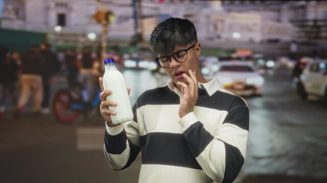 Man holding milk bottle and peering at the cap touches chin while inspecting label on a busy city street at night; doubt.