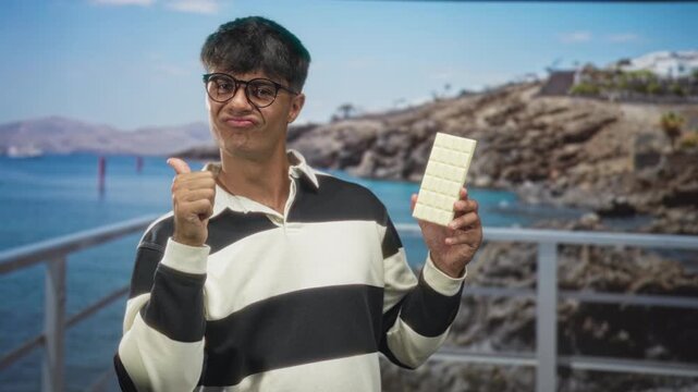Man holds white chocolate bar and points finger to chocolate in studio near seaside railing and rocks; playful craving.