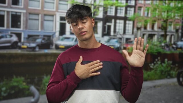 Young hispanic man with hand on chest and raised palm, touching and gesturing on city street; apology sincerity.