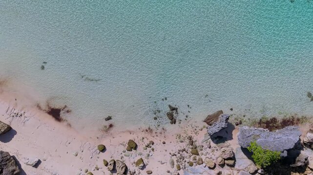 Top down aerial view of crystal clear turquoise sea water gently lapping a sandy tropical beach with coral reefs and coastal rocks