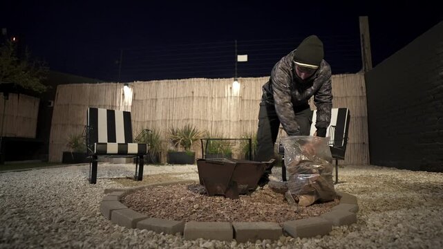 Person arranging firewood inside outdoor metal fire pit in residential backyard at night, showing fire preparation, gravel patio setting and casual cold weather leisure lifestyle, static wide shot.